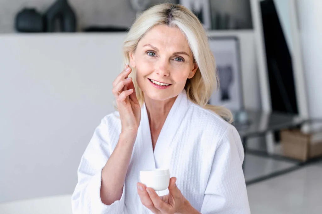 A smiling mature woman in a white bathrobe applying face cream.