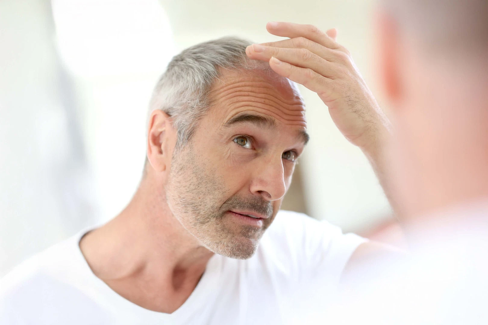 A mature man with gray hair and visible hair loss, looking into a mirror and running his fingers through his thinning hair.