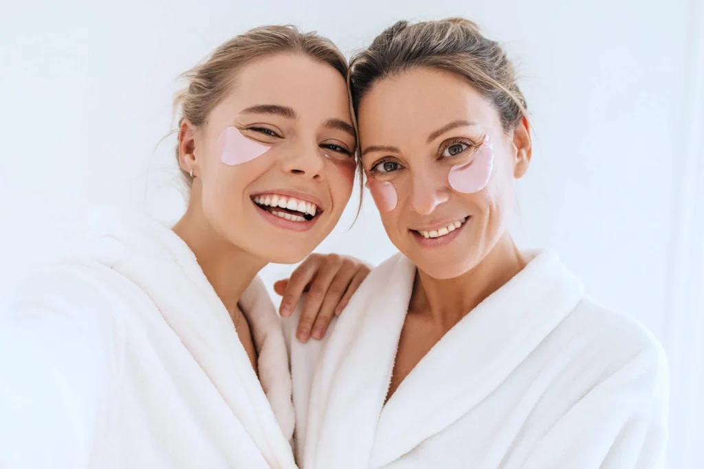 Two smiling women in white bathrobes wearing pink under-eye patches.